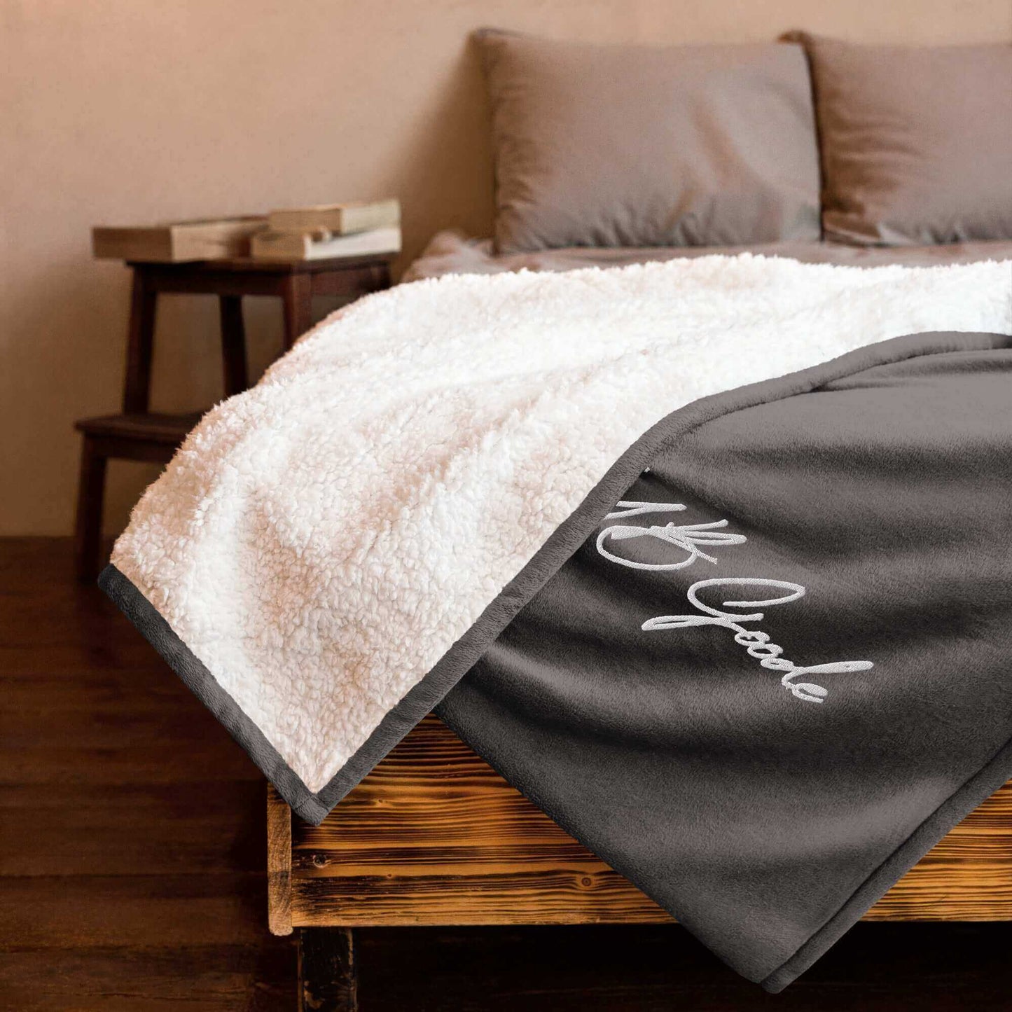 YB Goode signature embroidered sherpa blanket draped over a wooden bed with gray pillows, showcasing its soft texture.