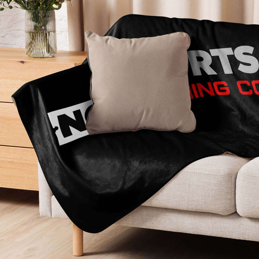 NO SHORTS CO. Sherpa Blanket on a sofa, featuring a smooth black fabric and a decorative pillow.
